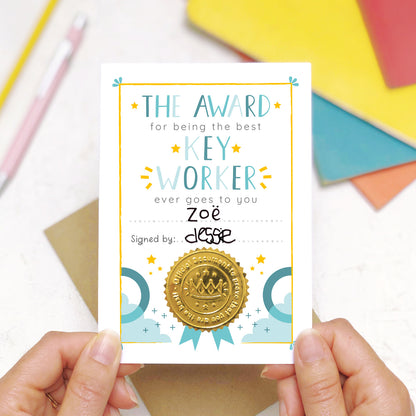 A nursery key worker award certificate card being held over a scene with a pen and pencil in the background along with some colourful exercise books. This is the blue version of the nursery key worker thank you card.