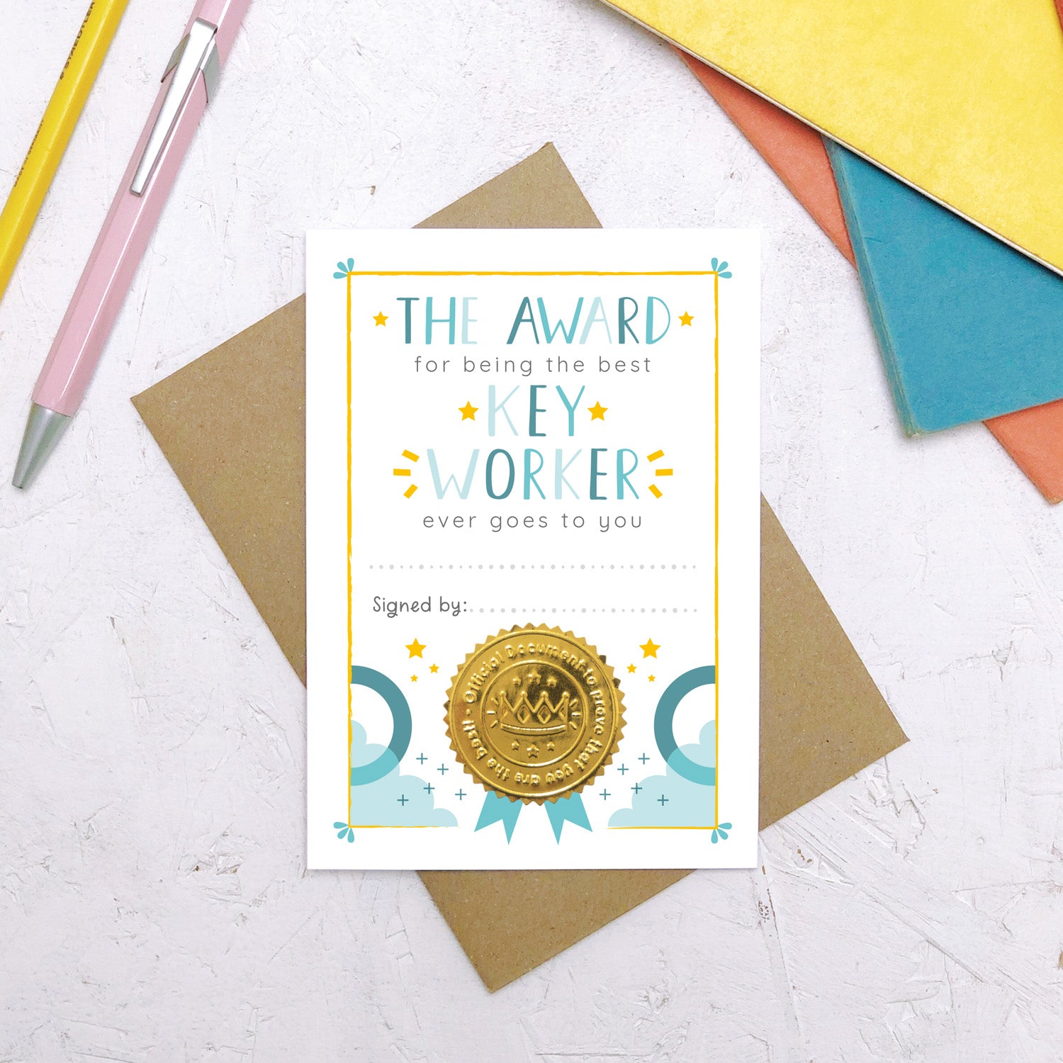 A nursery key worker award certificate card lying flat on top of a Kraft brown envelope surrounded by a pen, pencil and colourful exercise books. This is the blue version of the nursery key worker thank you card.