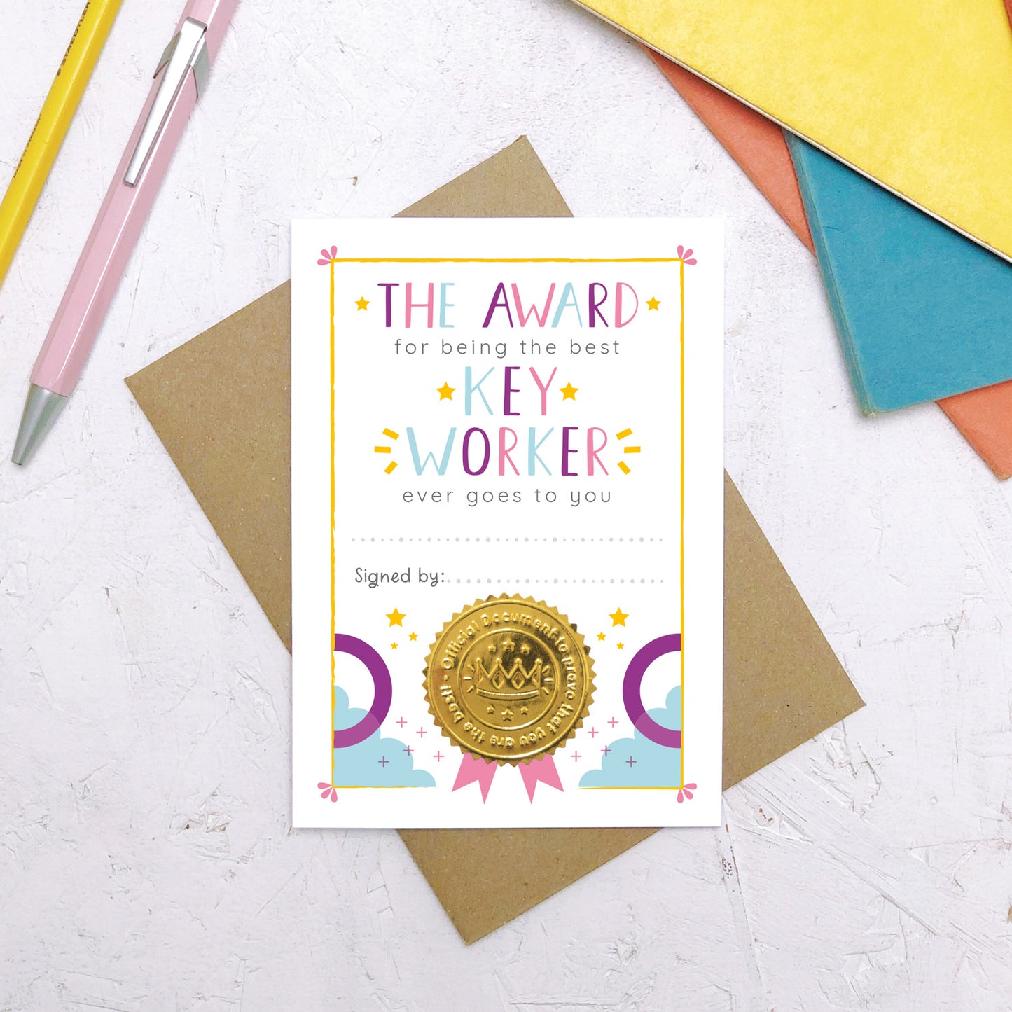 A nursery key worker award certificate card lying flat on top of a Kraft brown envelope surrounded by a pen, pencil and colourful exercise books. This is the pink, purple and blue version of the nursery key worker thank you card.