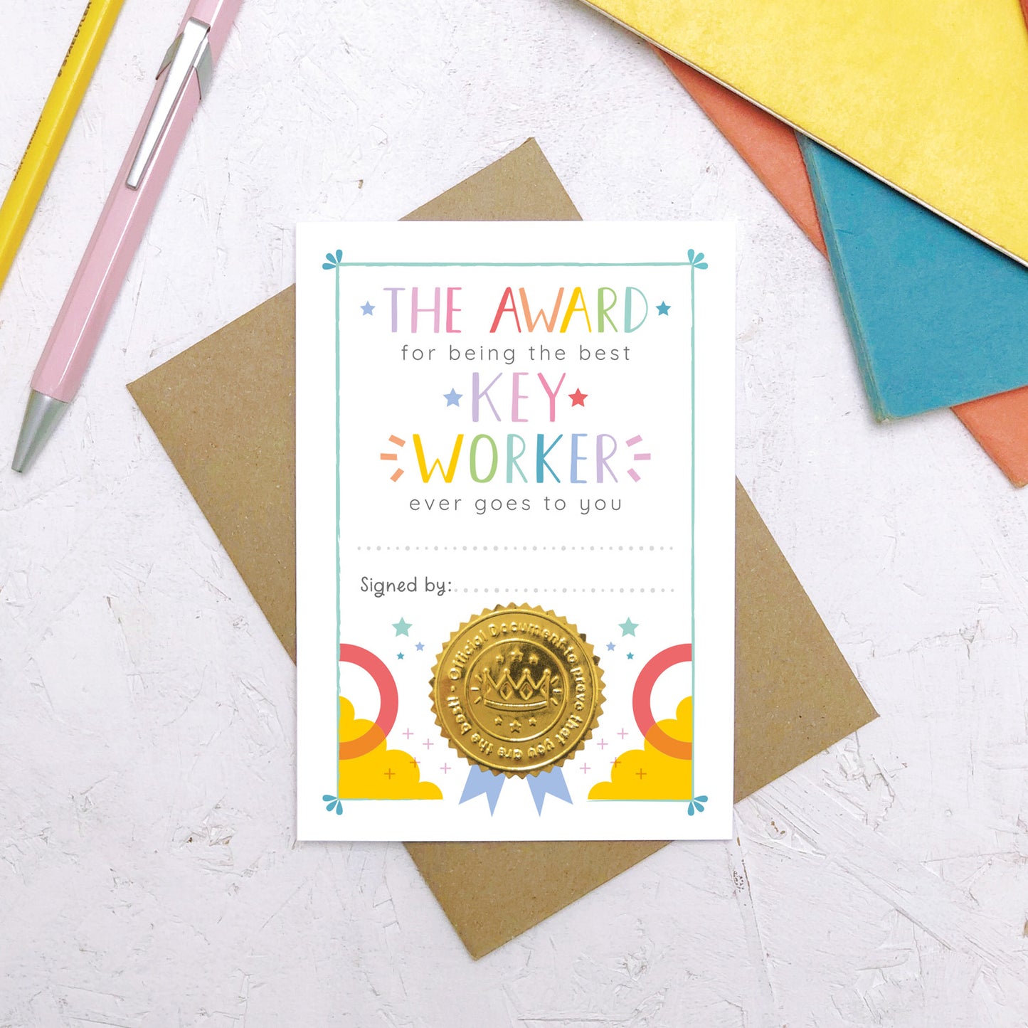 A nursery key worker award certificate card lying flat on top of a Kraft brown envelope surrounded by a pen, pencil and colourful exercise books. This is the rainbow version of the nursery key worker thank you card.