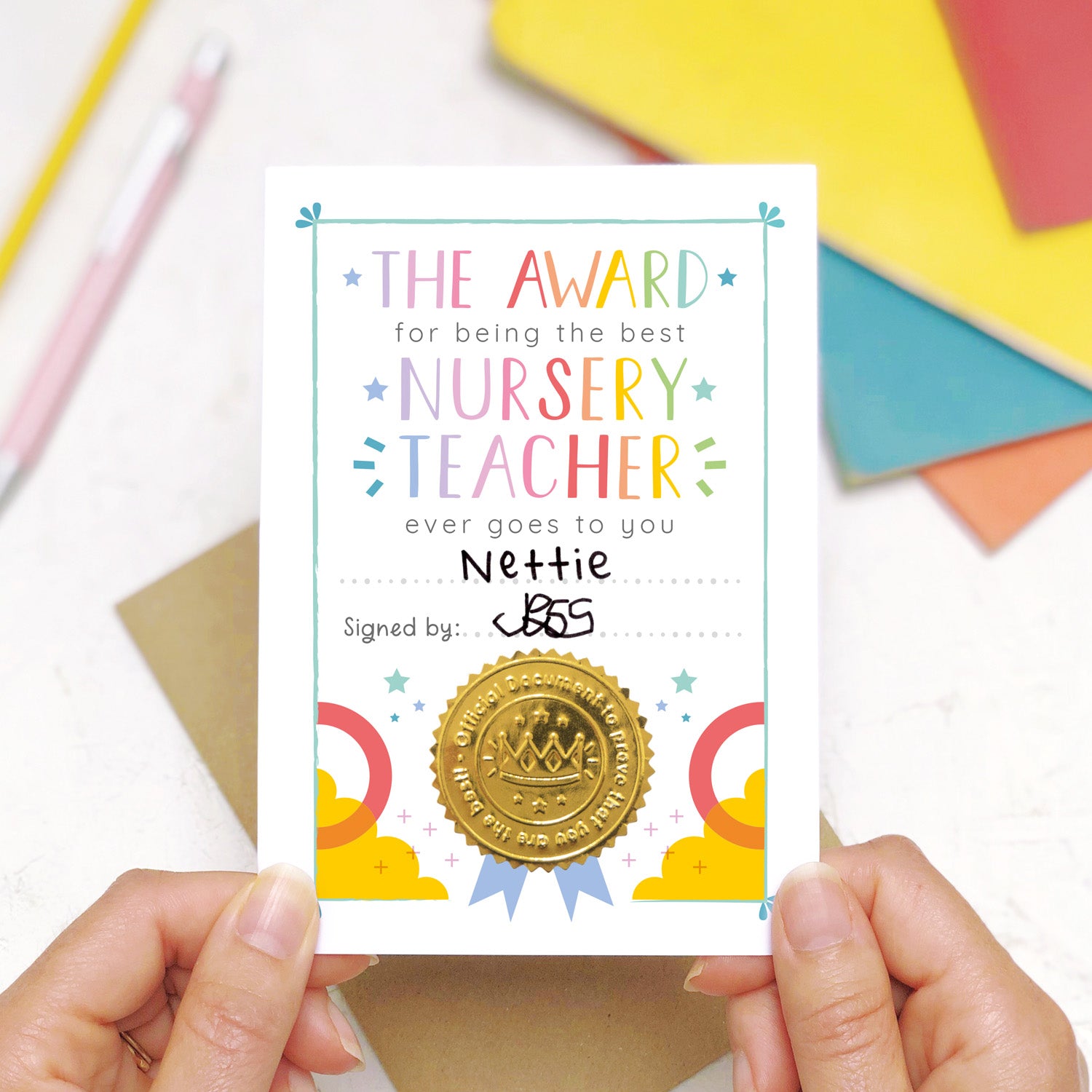 A nursery teacher award certificate card being held over a scene with a pen and pencil in the background along with some colourful exercise books. This is the rainbow version of the nursery teacher thank you card.