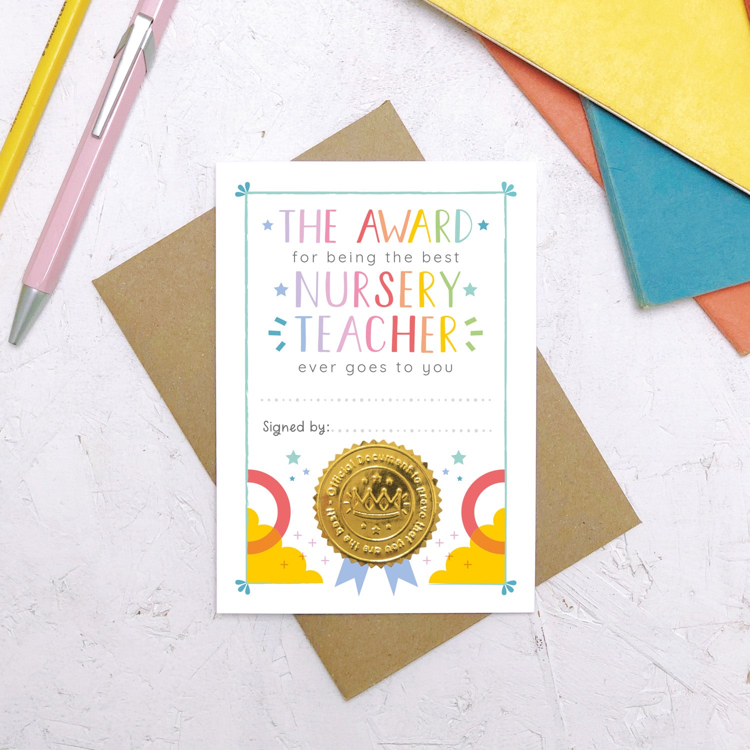 A nursery teacher award certificate card lying flat on top of a Kraft brown envelope surrounded by a pen, pencil and colourful exercise books. This is the rainbow version of the nursery teacher thank you card.