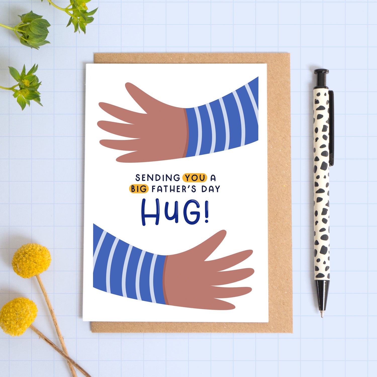 A card with two illustrated hands hugging the card and the phrase 'sending you a big father's day hug!'. This card is the version with the brown hands. The card is laid on a kraft brown envelope on top of a blue background next to a pen and flowers.