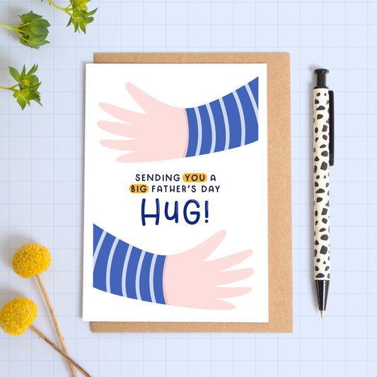 A card with two illustrated hands hugging the card and the phrase 'sending you a big father's day hug!'. This card is the version with the pink hands. The card is laid on a kraft brown envelope on top of a blue background next to a pen and flowers.