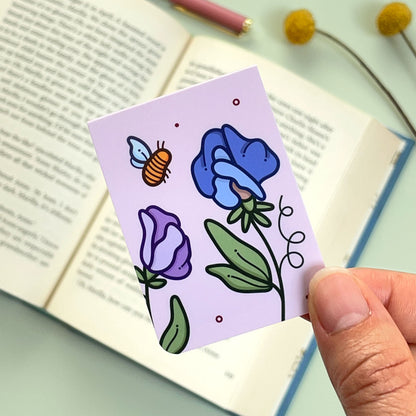 A floral sweet pea bookmark with purple and blue flowers and a bee held over an open book