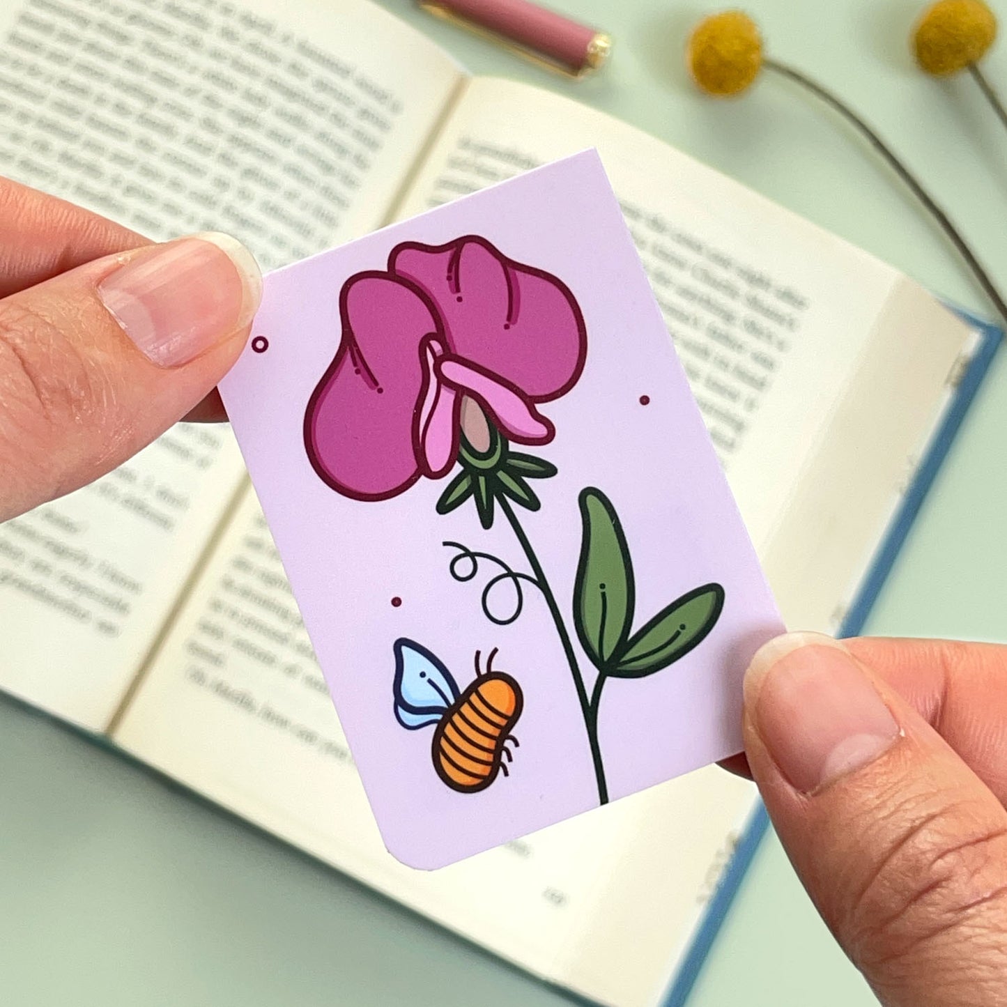 The reverse side of the floral sweet pea bookmark with a pink flower and a bee held over an open book