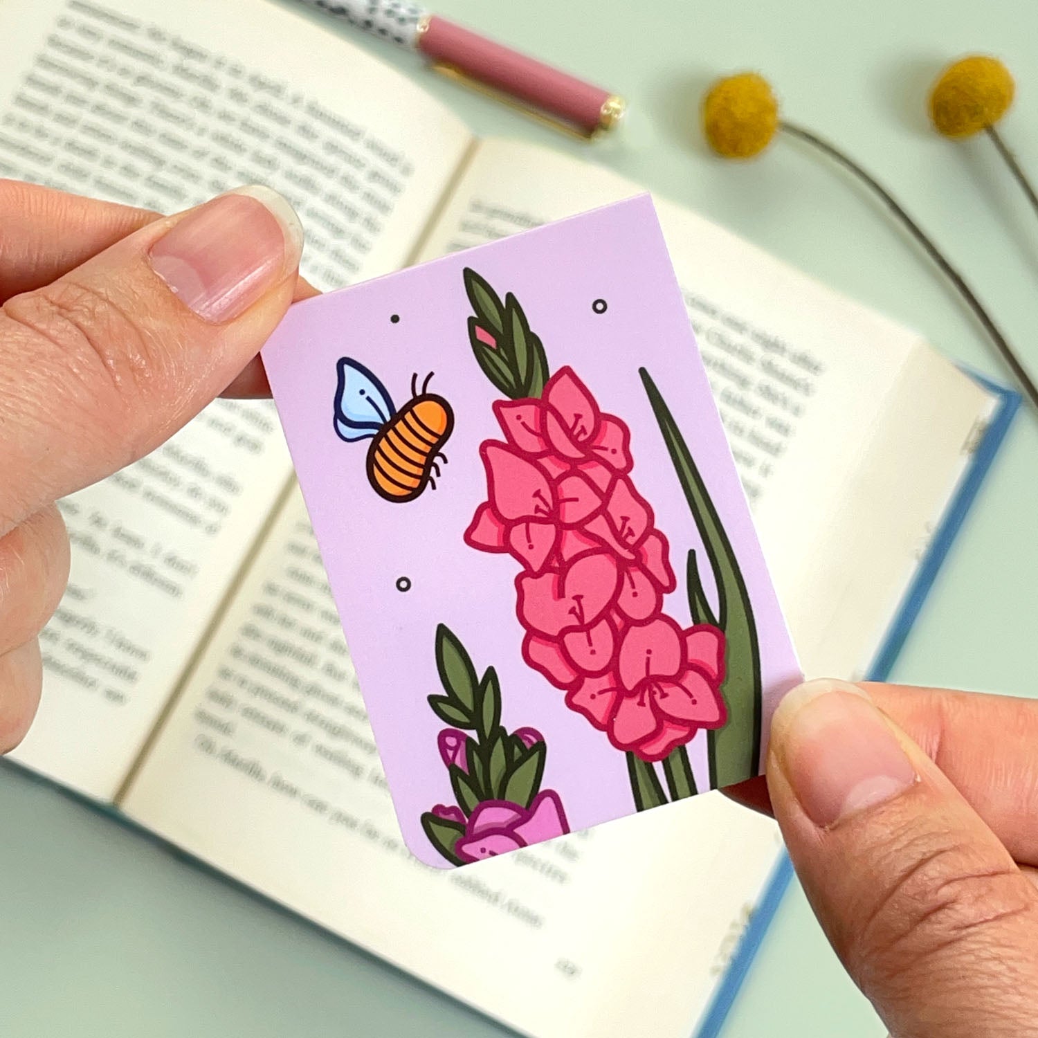 The reverse side of the floral gladiolus bookmark with red and pink flowers and a bee held over an open book