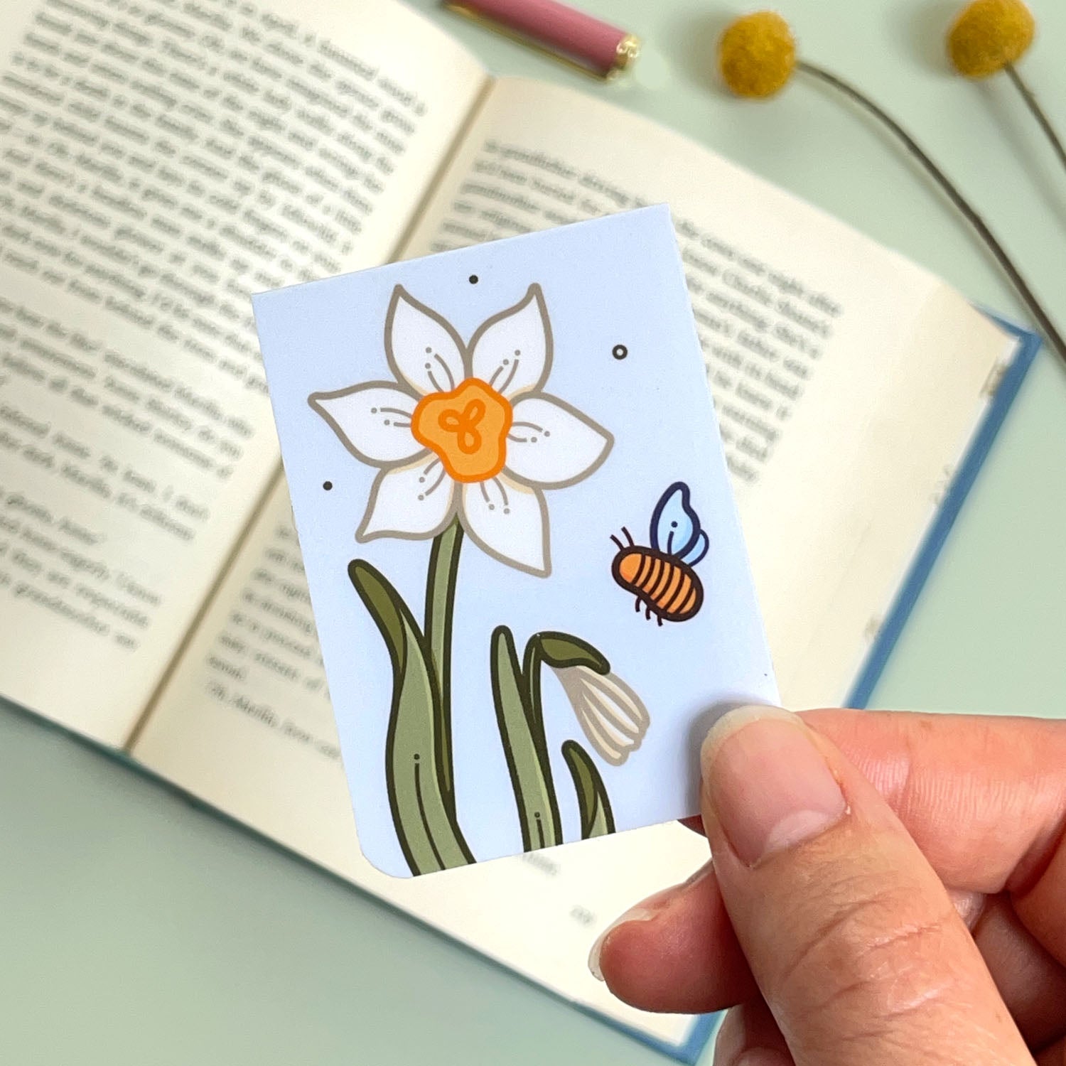 A floral narcissus bookmark with cream flowers and a bee held over an open book