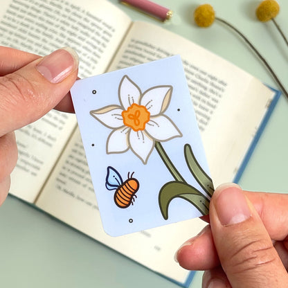 The reverse side of the floral narcissus bookmark with cream flowers and a bee held over an open book