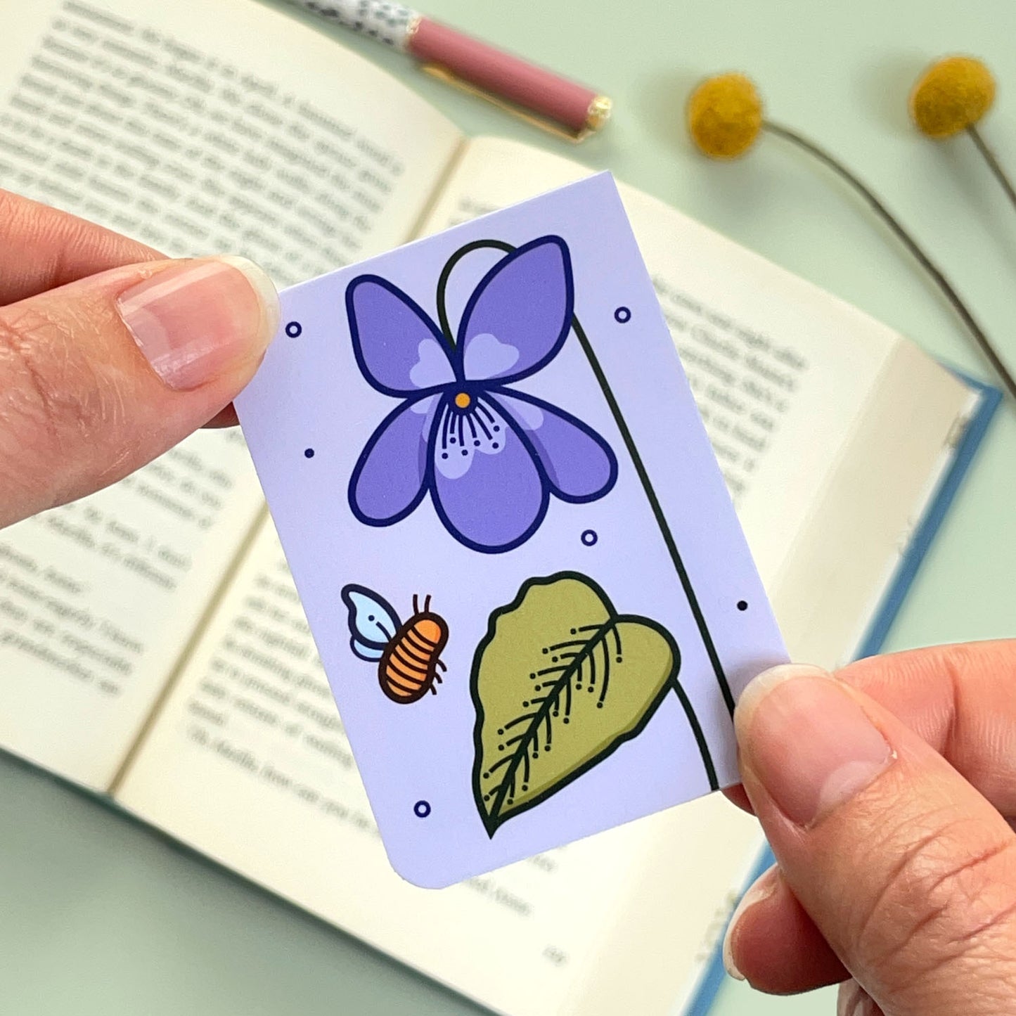 A floral violet bookmark with purple flowers and a bee held over an open book