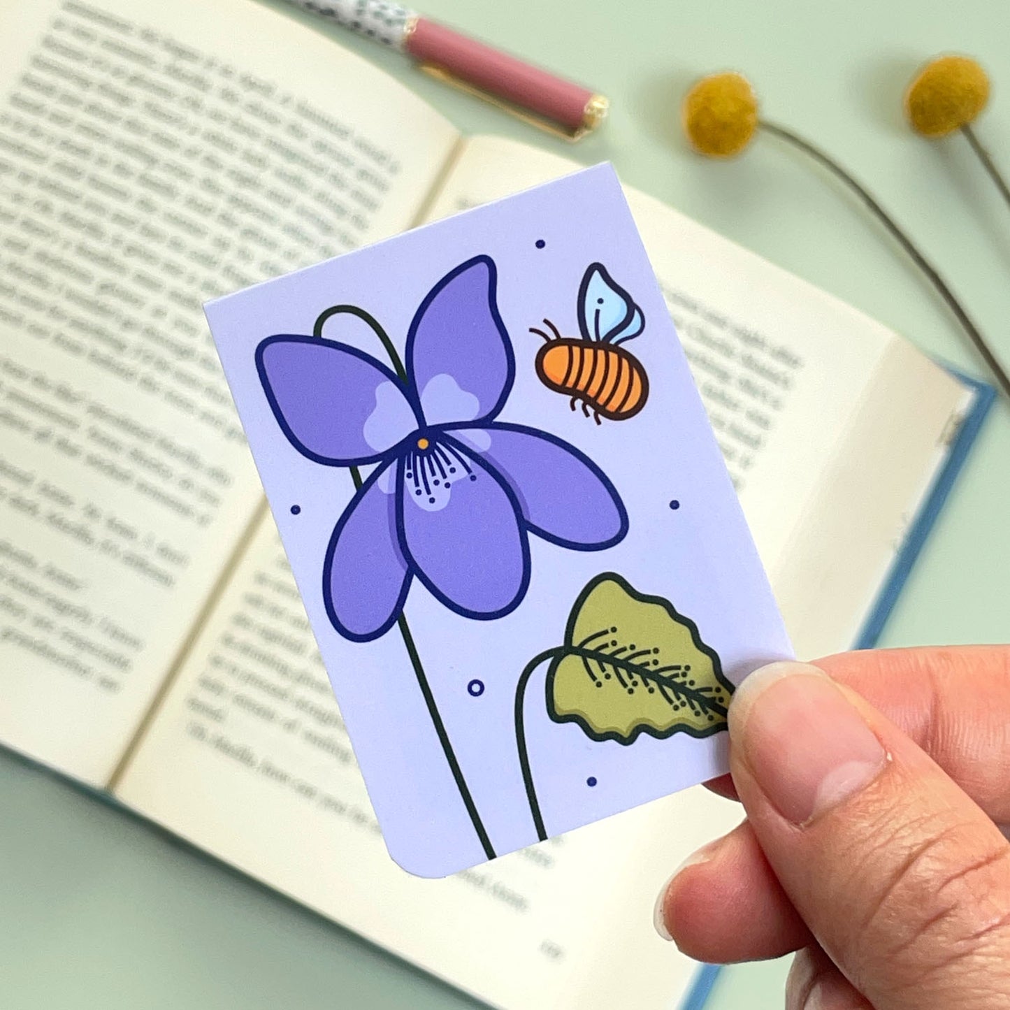 The reverse side of the floral violet bookmark with violet flowers and a bee held over an open book