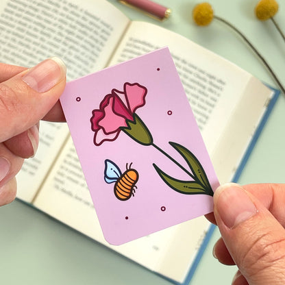 The alternate side of the floral carnation bookmark with pink flowers and a bee held over an open book