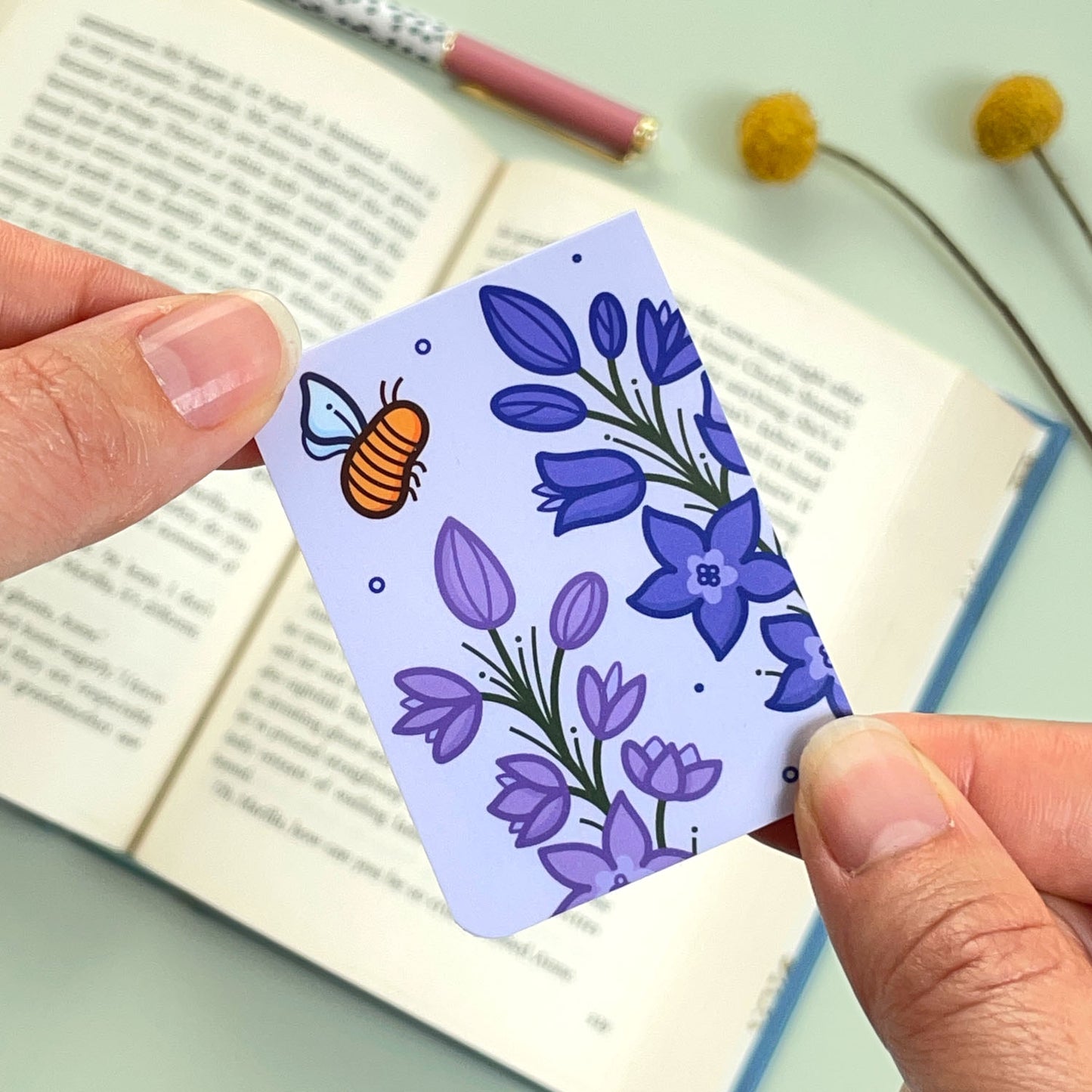 A floral Larkspur bookmark with purple flowers and a bee held over an open book