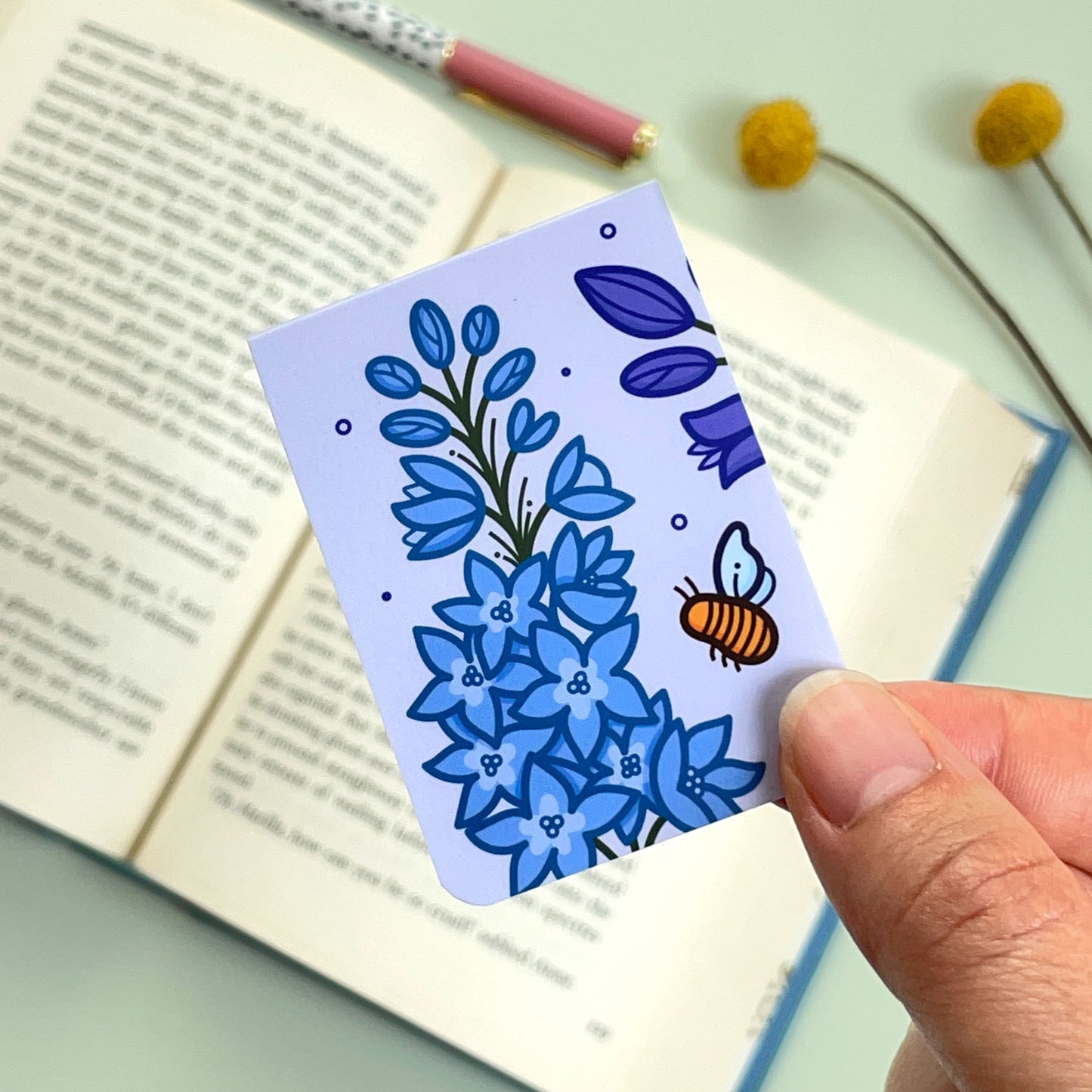 The reverse side of the floral Larkspur bookmark with a purple and blue flowers and a bee held over an open book