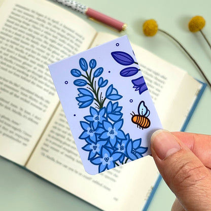 The reverse side of the floral Larkspur bookmark with a purple and blue flowers and a bee held over an open book