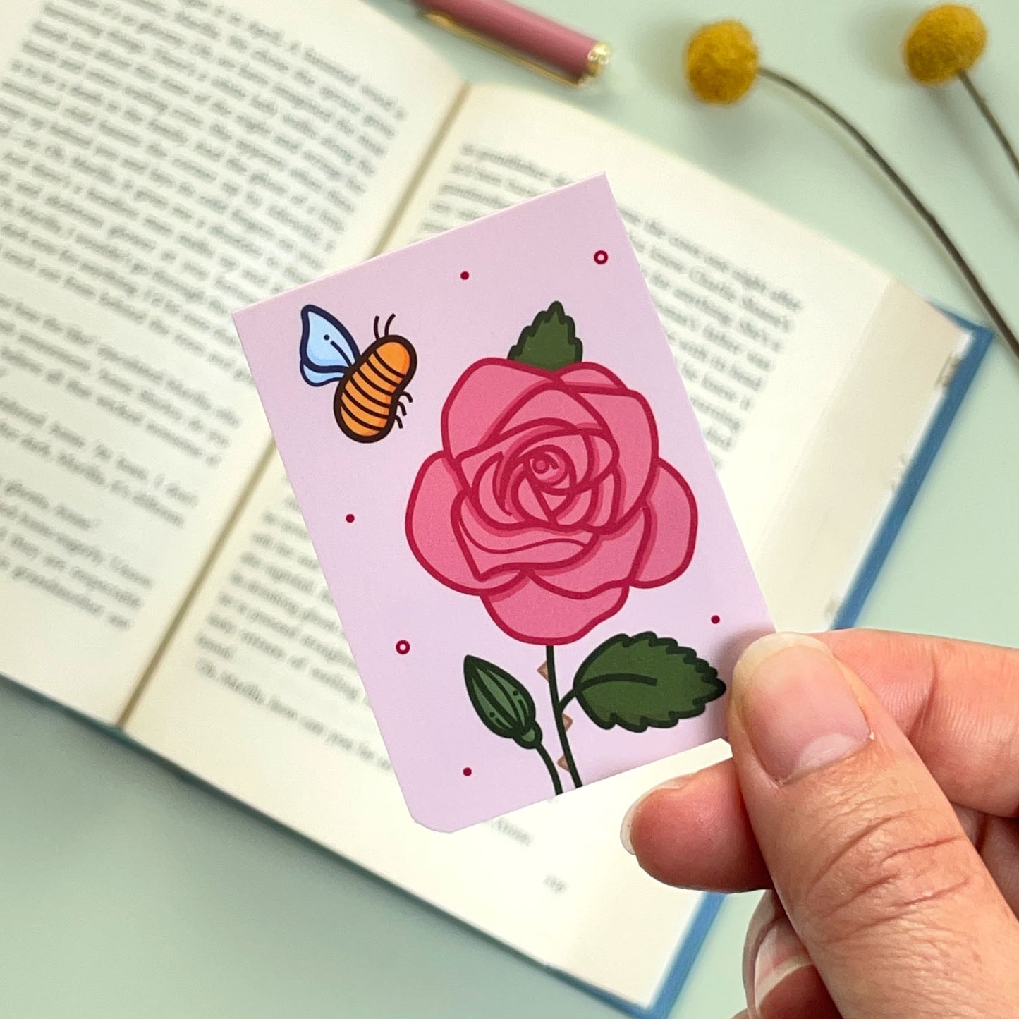 A floral rose bookmark with green leaves and pink flowers and a bee held over an open book