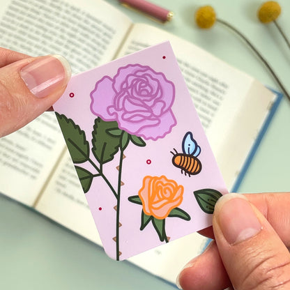 The reverse side of the floral rose bookmark with a pink and peach flower and a bee held over an open book