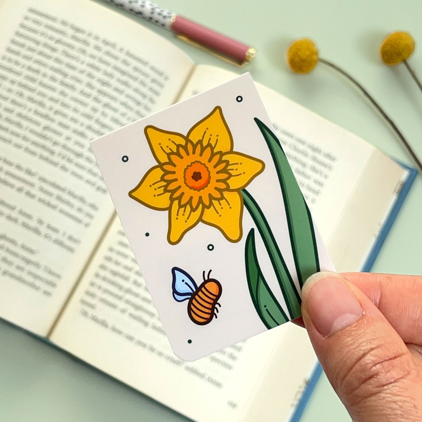 A floral daffodil bookmark with a yellow flower and a bee held over an open book