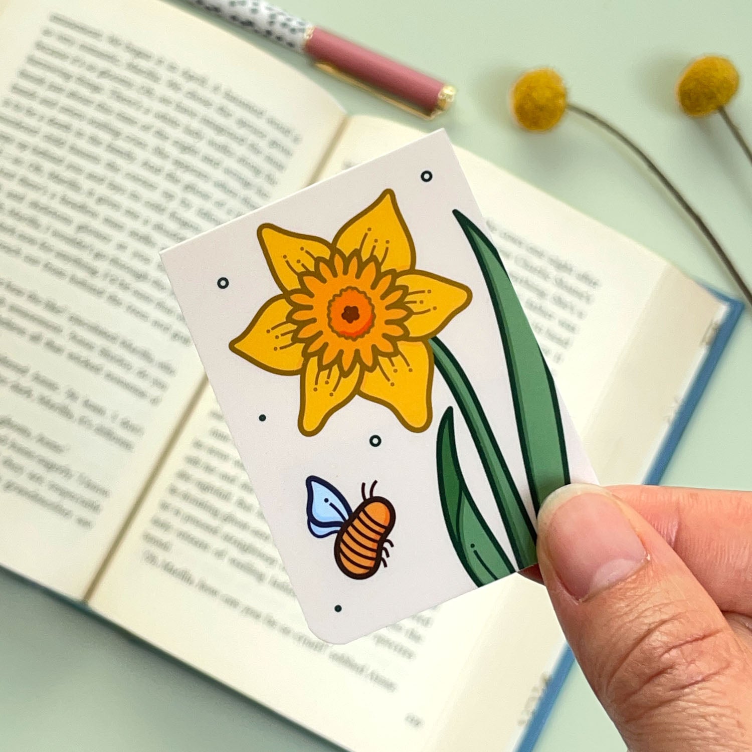 A floral daffodil bookmark with a yellow flower and a bee held over an open book