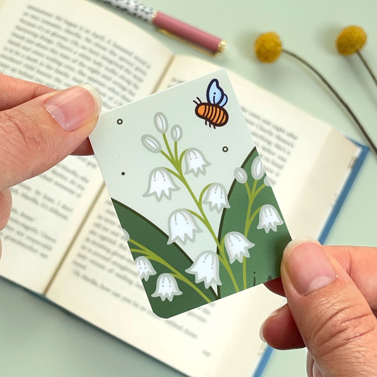 A floral lily of the valley bookmark with green leaves and cream flowers and a bee held over an open book