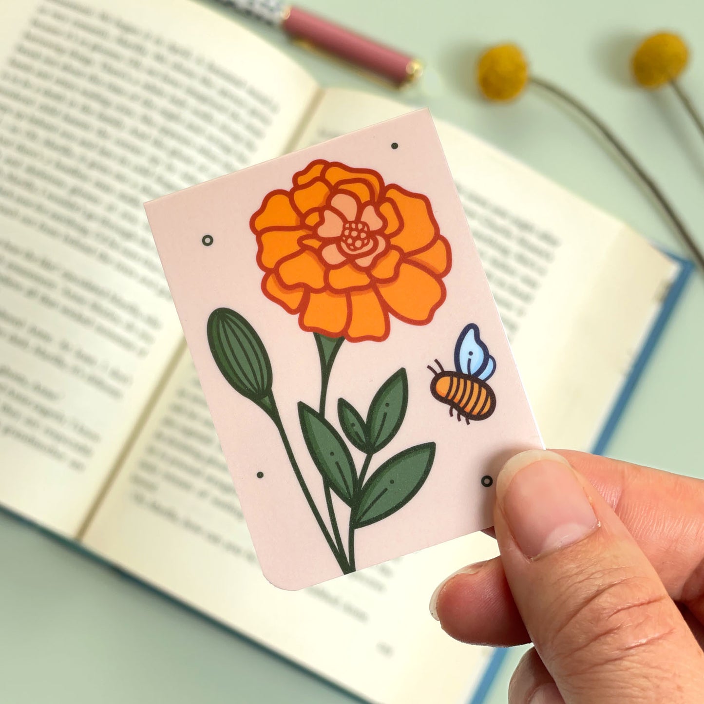 A floral marigold bookmark with orange flowers and a bee held over an open book