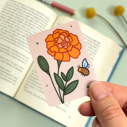 A floral marigold bookmark with orange flowers and a bee held over an open book