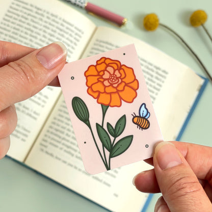 The reverse side of the floral marigold bookmark with orange flowers and a bee held over an open book