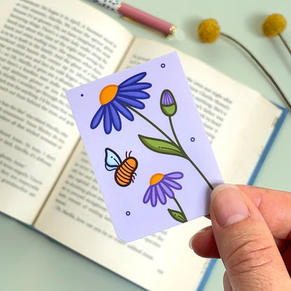 A floral aster bookmark with purple flowers and a bee held over an open book