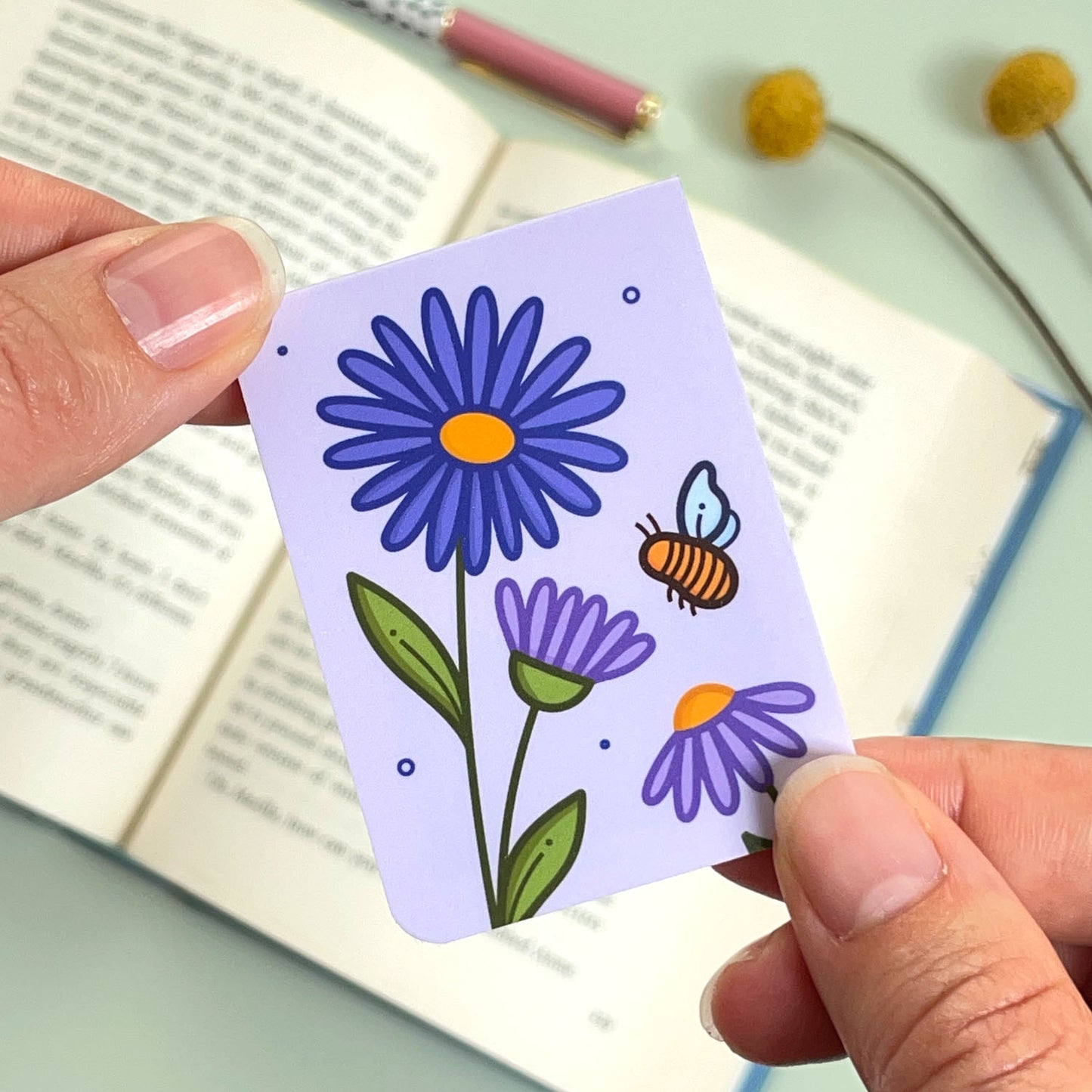 The reverse side of the floral aster bookmark with purple flowers and a bee held over an open book