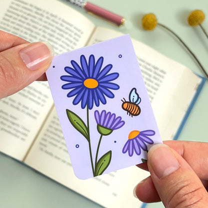 The reverse side of the floral aster bookmark with purple flowers and a bee held over an open book