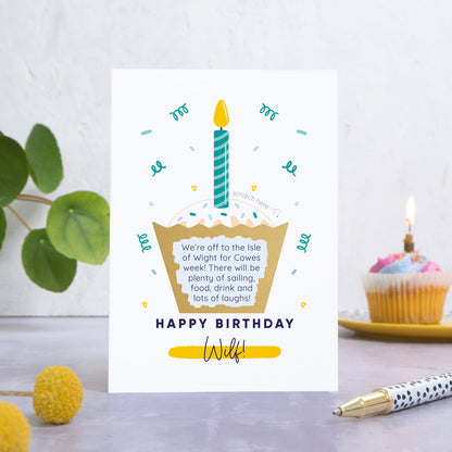 A birthday cake scratch card featuring an illustration of a cup cake with a candle in the top. The wording underneath the cake reads 'happy birthday' and then the name of the recipient is printed underneath. This card has been photographed on a grey back drop with leaves and a birthday cake in the background. The cakes wrapper has been scratched off to reveal the personalised message.  This is the blue card colourway.