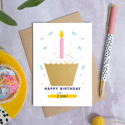 The birthday cake scratch card lying down top of a kraft brown envelope on a grey background surrounded by leaves, a pen and a small cupcake. The card features a cupcake with a pink candle, confetti and still has the gold scratch panel unscratched.