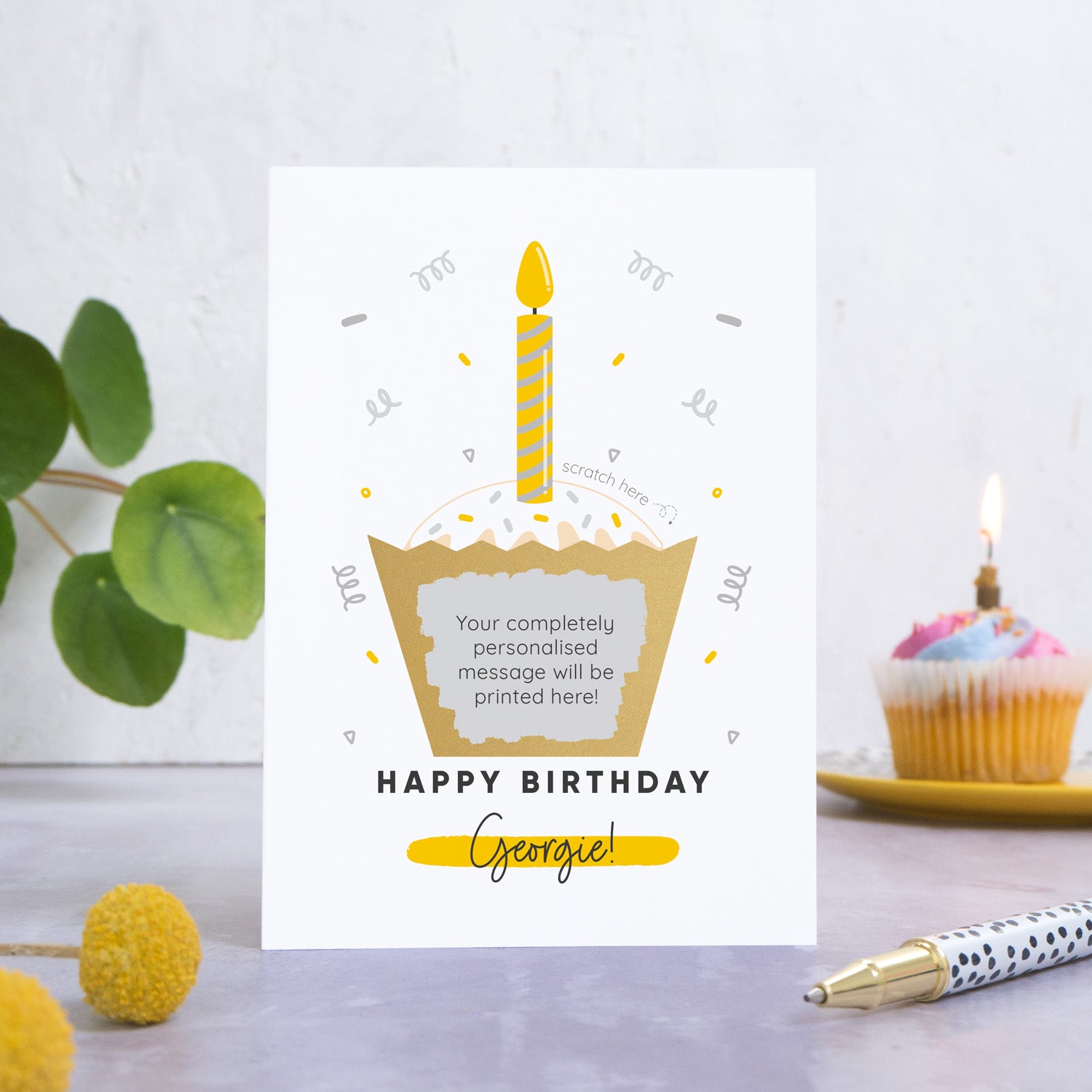 A birthday cake scratch card featuring an illustration of a cup cake with a candle in the top. The wording underneath the cake reads 'happy birthday' and then the name of the recipient is printed underneath. This card has been photographed on a grey back drop with leaves and a birthday cake in the background. The cakes wrapper has been scratched off to reveal the personalised message.  This is the grey card colourway.