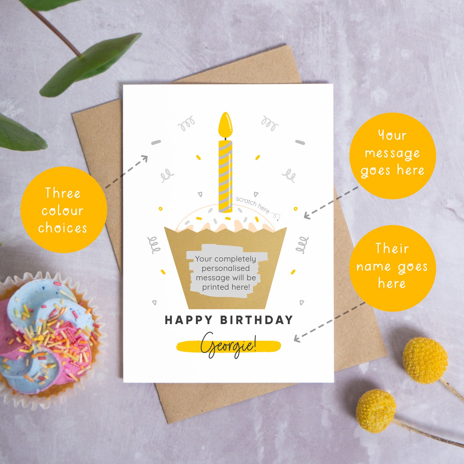The birthday cake scratch card lying down top of a kraft brown envelope on a grey background surrounded by leaves, a pen and circles explaining the various points of that card that can be personalised. The card features a cupcake with a yellow and grey candle, confetti and still has the gold scratch panel has been scratched to reveal the secret message.