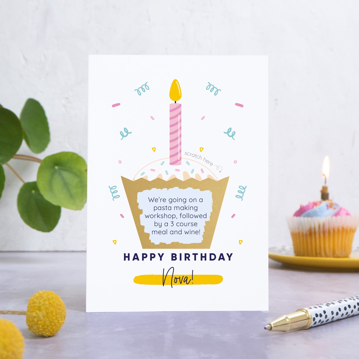A birthday cake scratch card featuring an illustration of a cup cake with a candle in the top. The wording underneath the cake reads 'happy birthday' and then the name of the recipient is printed underneath. This card has been photographed on a grey back drop with leaves and a birthday cake in the background. The cakes wrapper has been scratched off to reveal the personalised message.  This is the pink card colourway.