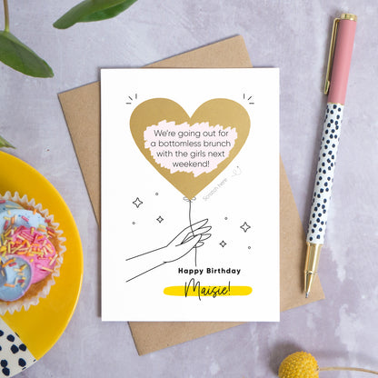 A personalised heart balloon birthday scratch card which has been photographed lying on a kraft brown envelope on top of a grey back drop with leaves, a pen and a cake in the background. The card features a gold scratch off balloon being held by an outlined hand with the wording beneath reading 'happy birthday Maisie!' 
