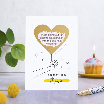 A personalised heart balloon birthday scratch card which has been photographed on a grey back drop with leaves and a cake in the background. The card features a gold scratch off balloon being held by an outlined hand with the wording beneath reading 'happy birthday Maisie!' 
