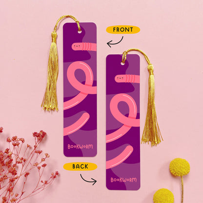 An image depicting the front and the back of the double sided metal bookmark which has been photographed on a pink background with dried flowers. Each bookmark has a gold tassel. The front and the back of the bookmark are printed with the same design of a happy pink worm.