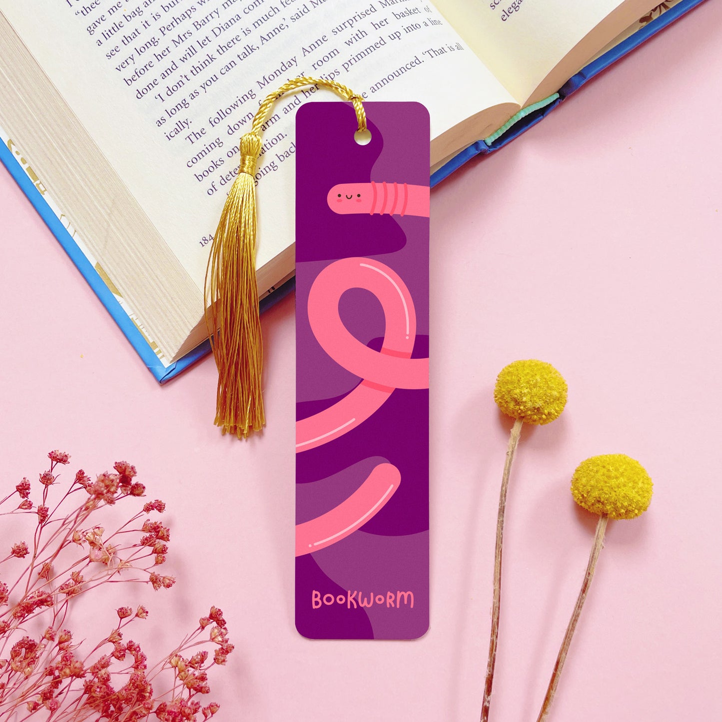 A bookworm themed, double sided, aluminium bookmark with a gold tassel which has been photographed on a pink background with an open book and dried flowers.