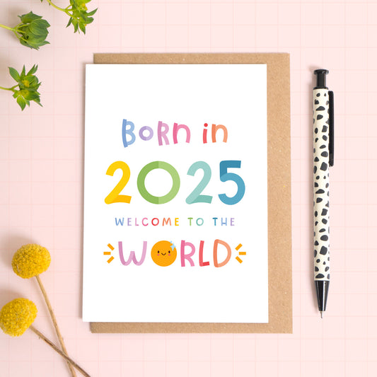 The unisex 'born in 2025' new baby card has been photographed lying on top of a Kraft brown envelope on a pink background. To the left is foliage and flowers and to the right is a black and white pen. The text reads: 'born in 2025 welcome to the world'.
