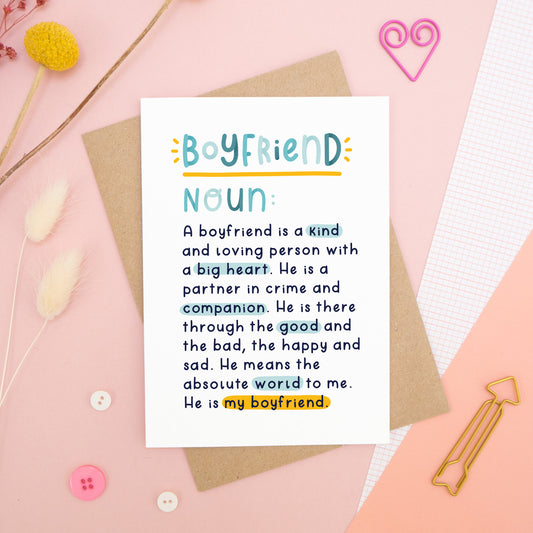 A heartfelt, colourful dictionary definition style card with the definition of a boyfriend, photographed on a pink background with floral props, paper clips, and buttons. 