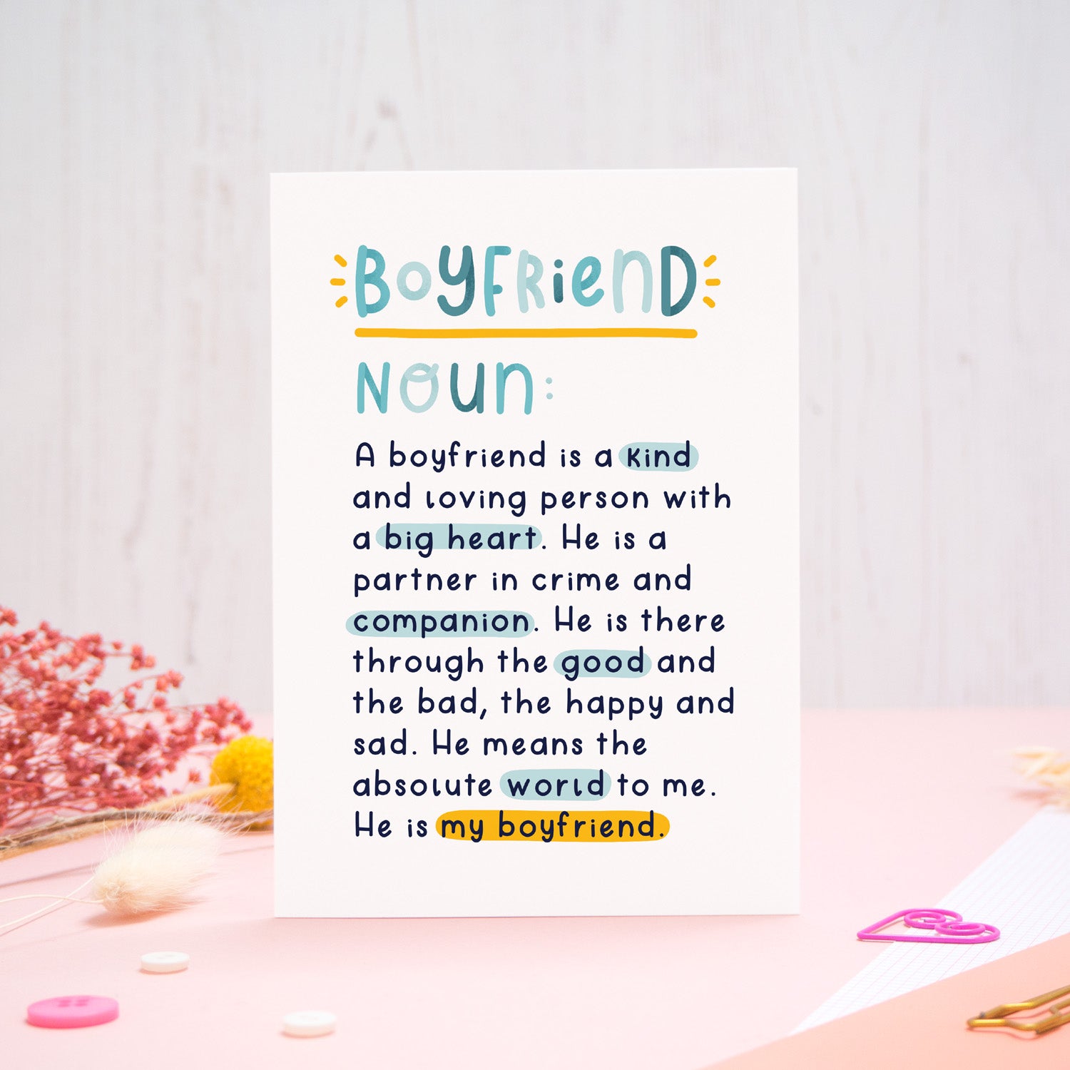 A heartfelt, colourful dictionary definition style card with the definition of a boyfriend, photographed stood on a pink and white background with floral props, paper clips, and buttons. 