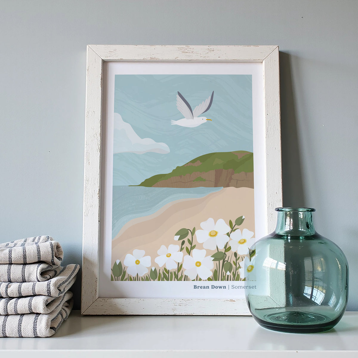 A framed coastal art print featuring a flying seagull, beach, flowers, and the green cliffs of Brean Down, styled on a shelf beside folded striped towels and a translucent blue glass bottle against a light grey wall.