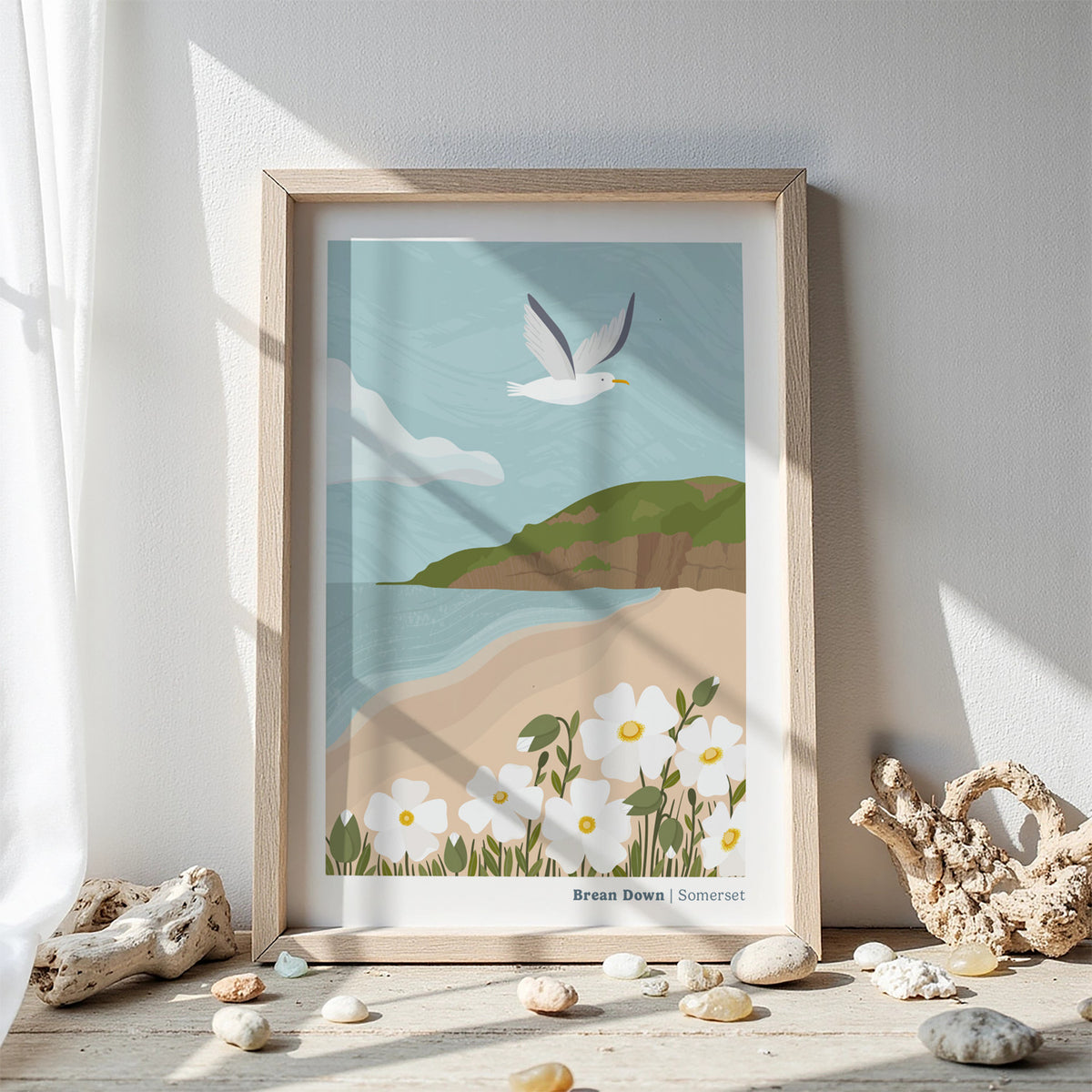 A framed art print of Brean Down in Somerset' featuring an illustration of the calm coastal landscape with a flying seagull, and white flowers displayed on a wooden surface with decorative driftwood and pebbles against a white wall.