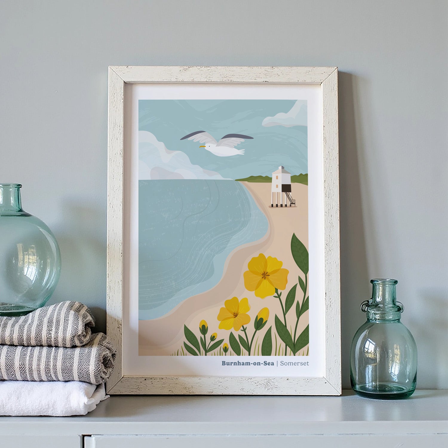 A coastal art print of the Low Lighthouse at Burnham-on-Sea inside of a white picture frame next to two blue bottles and a pile of linen cloths.