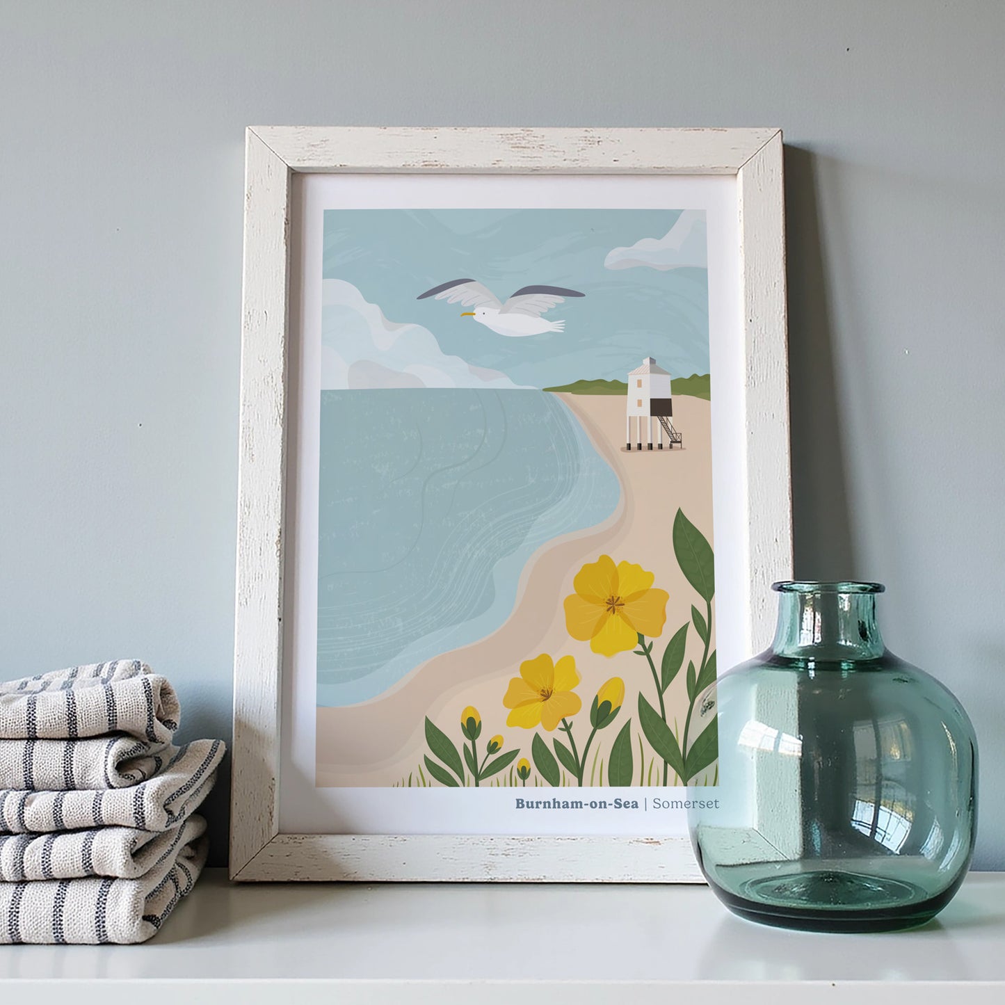 A coastal art print of the Low Lighthouse at Burnham-on-Sea inside of a white picture frame behind some linen cloths and a blue vase.