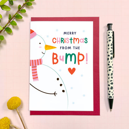 A snowman themed 'from the baby bump' Christmas card photographed on a red envelope on top of a pink background surrounded by leaves, yellow flowers and a pen for scale.