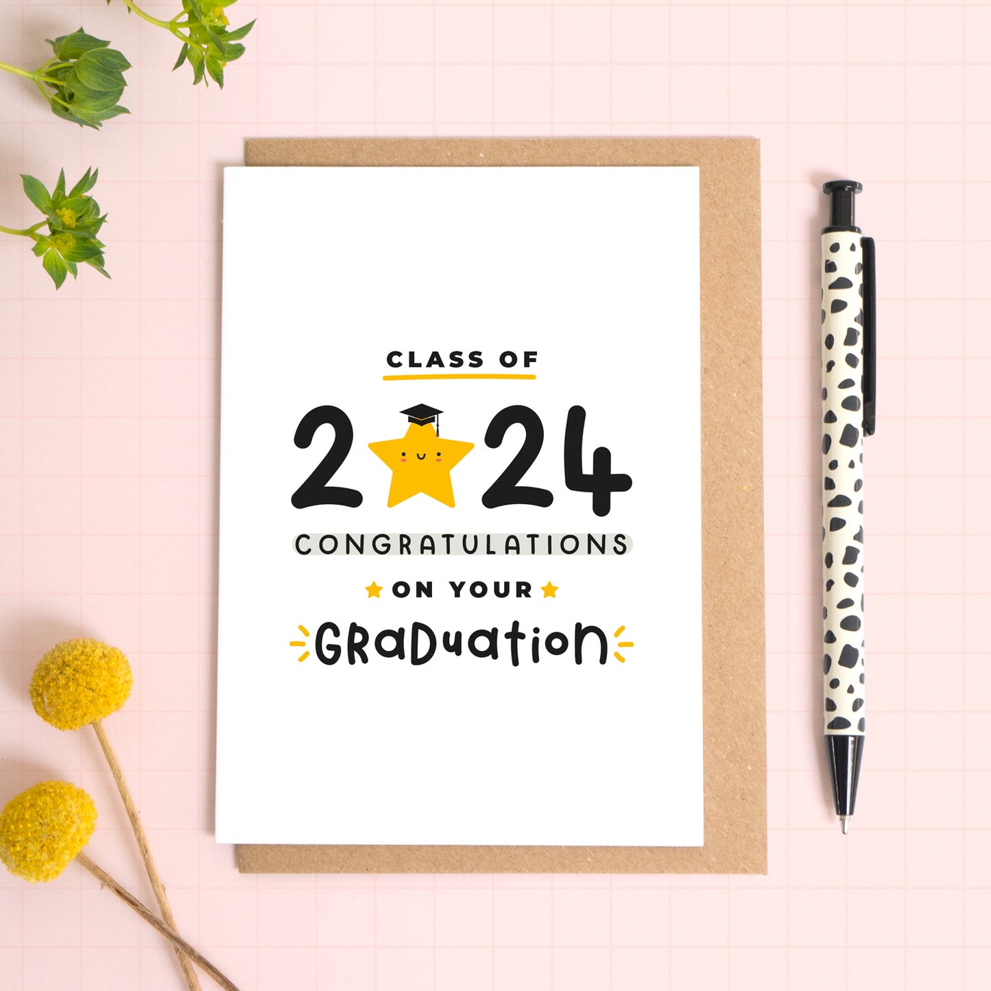 A class of 2024 graduation card. This card has been shot lying down on a kraft brown envelope and there is foliage to the side and a pen for scale. The text on this card reads 'class of 2024 congratulations on your graduation.