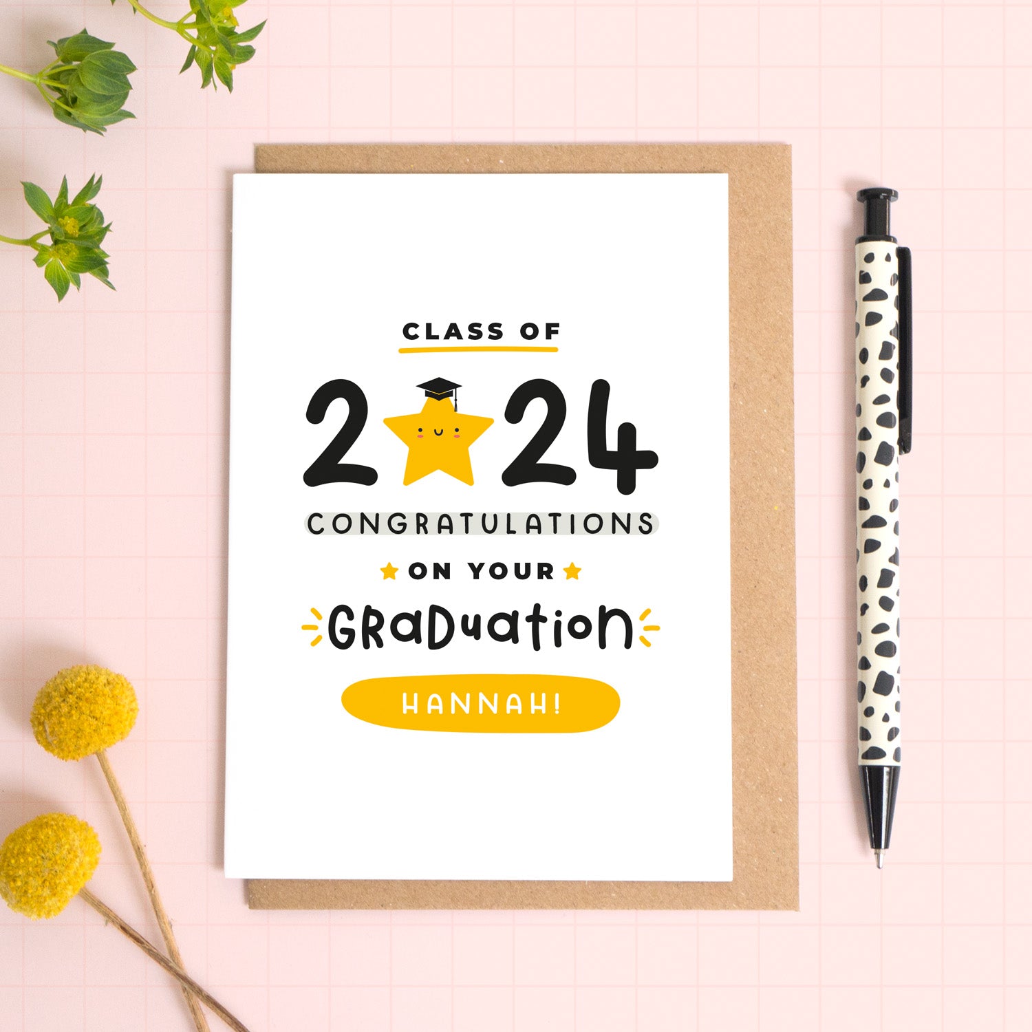 A personalised class of 2024 card complete with the recipients name and class year. This card has been shot lying down on a kraft brown envelope and there is foliage to the side and a pen for scale. The text on this card reads 'class of 2024 congratulations on your graduation hannah!'