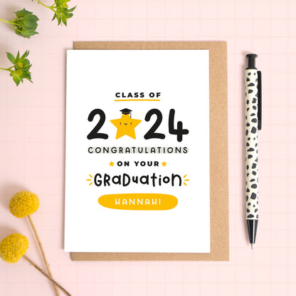 A personalised class of 2024 card complete with the recipients name and class year. This card has been shot lying down on a kraft brown envelope and there is foliage to the side and a pen for scale. The text on this card reads 'class of 2024 congratulations on your graduation hannah!'